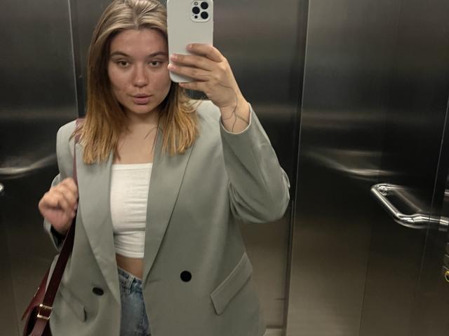 LioraEm with blonde hair, wearing a grey blazer, white tube top, and blue jeans, taking a selfie in a metal elevator, holding a brown bag in her other hand.