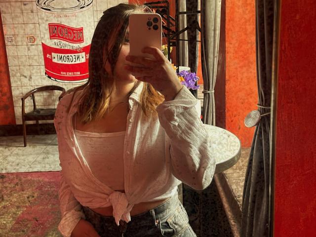 LioraEm with medium tits, wearing a tied-up white shirt and blue jeans, taking a mirror selfie with an iPhone, covering her face. Red-orange walls in the background.