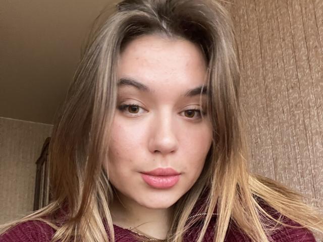 LioraEm has light brown eyes, straight blonde hair with darker roots, and pink lipstick. She's wearing a maroon top, with a subtle, cute expression.