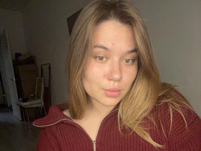LioraEm has light brown hair, brown eyes, and is making a pouty face with slightly parted lips. She's wearing a red zip-up sweater. Her ass is not visible.