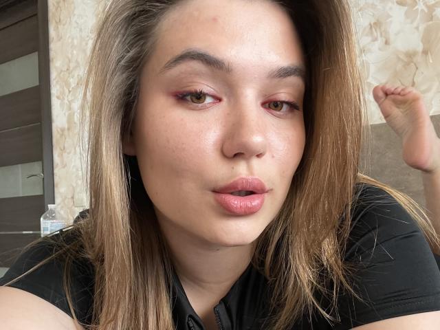 LioraEm has shiny skin, pouty pink lips, and brown eyes with red eyeshadow. She's sucking her bottom lip, wearing a black shirt, with dirty blonde hair.