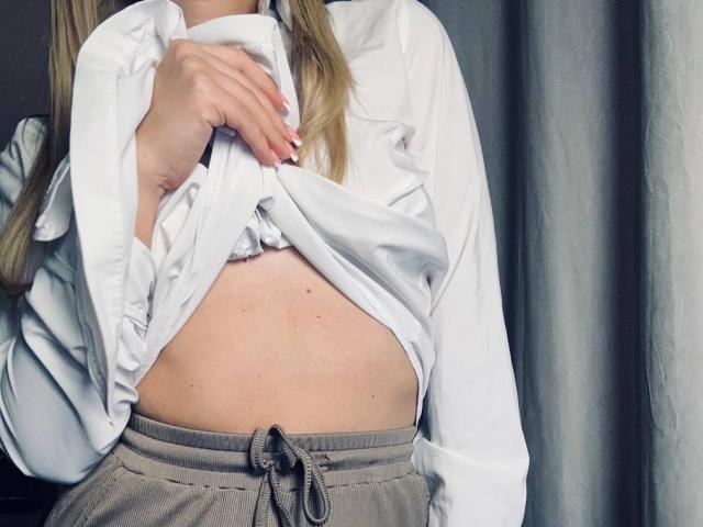 BalletSwan lifting a white shirt to reveal her bare, soft, slightly freckled belly, wearing gray pants with a drawstring, blonde hair visible, hand with pink nails on shirt.