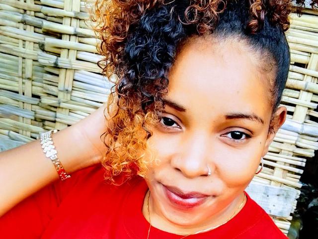 MiriamX69 with curly brown and orange hair, red shirt, nose ring, hoop earrings, silver bracelet, smiling at camera, hand behind head, tan skin, cute, attractive.