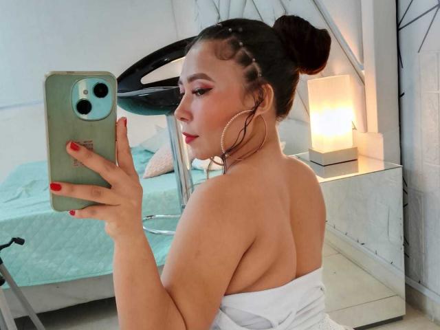 JadeFoxx, con el cabello trenzado en un moño, grandes aros, uñas rojas, tomándose un selfie, mostrando su espalda desnuda y una toalla blanca, sosteniendo un teléfono verde..