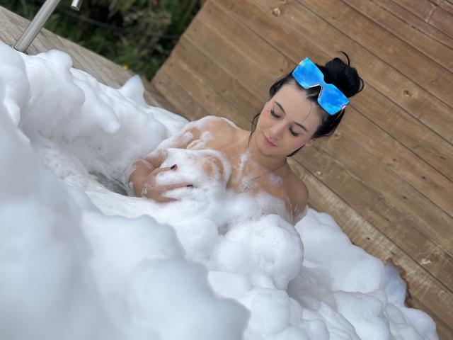 MishuHottie in a bubble-filled hot tub, wearing blue sunglasses on her black hair, with soapy white bubbles covering her tits and upper body. Wood panel wall in background.