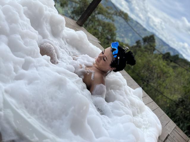 MishuHottie with black hair in a blue hairpin, sitting in a wooden hot tub filled with white foam, her tits partially covered, eyes closed, in a forested area.