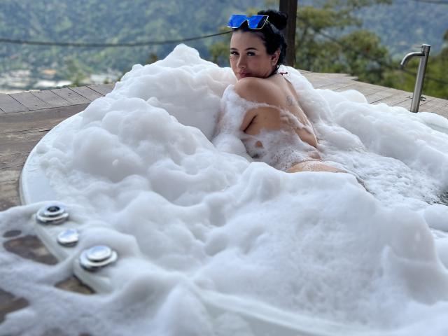 MishuHottie in a hot tub, covered in white bubbles, looking back with blue sunglasses on her head, showing her ass and back, surrounded by wood and metal.