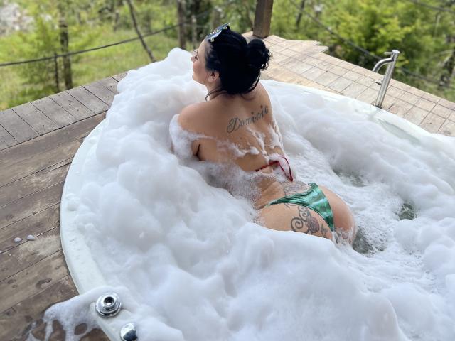 MishuHottie in a hot tub, back to camera, bubbles covering her, green bikini bottom, tattoos on back and ass, black hair in bun, wooden deck, forest in background.