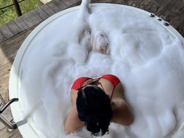 MishuHottie with black hair, red polka-dot bikini top, and tan skin, lying face-down in a white bubble bath, her ass and back visible, in a round wooden tub.