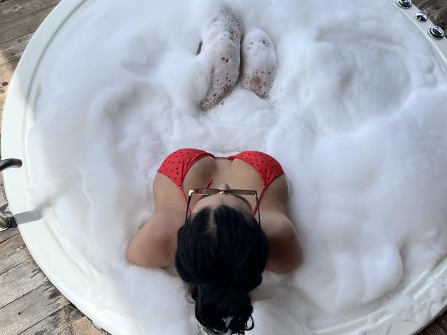 MishuHottie in a red bikini, lying face down in a white, foamy tub. Her black hair is in a bun, and her ass is partially covered by bubbles.