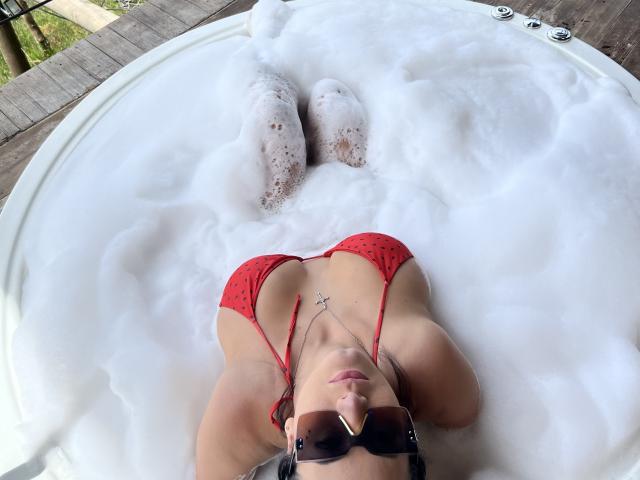 MishuHottie with red polka-dot bikini, small tits, lying in a bubble bath, wearing sunglasses, legs up, white foam covering, necklace with cross, wooden deck outside.