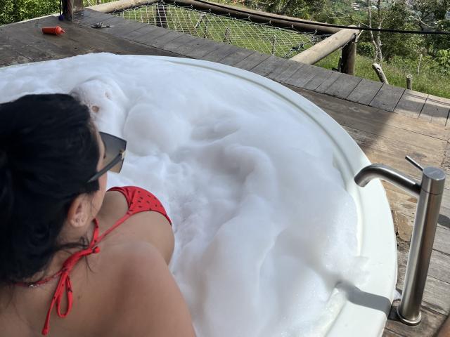 MishuHottie with black hair, red bikini top, and sunglasses, soaking in a white, bubbly hot tub, showing her back and upper ass, on a wooden deck.
