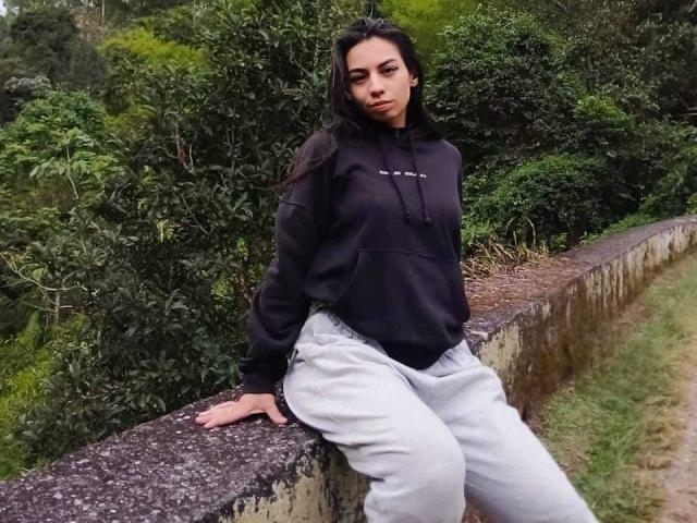 KatherinJohns with dark hair, black hoodie, and gray pants, sitting on a mossy stone wall, arms resting, looking slightly annoyed, in a green, wooded area.