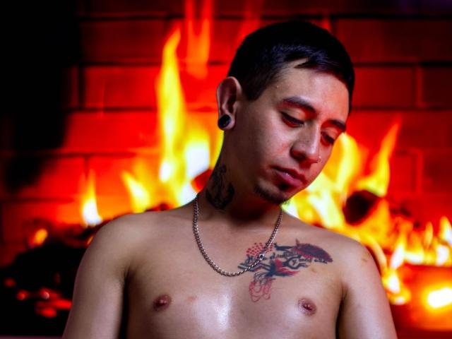 Abnedarm with short black hair, red eye makeup, and a chain necklace, shirtless, showing tattoos on chest and neck, standing in front of a bright red fire.