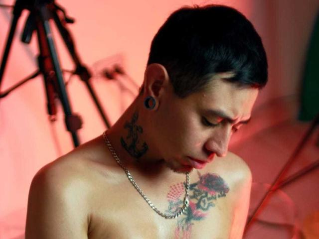 Abnedarm with short black hair, tattoos on neck and chest, large black ear plugs, and a chain necklace, looking down with a serious expression, topless.