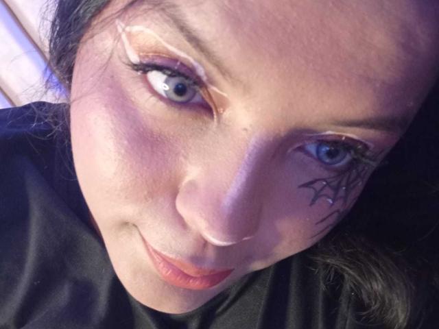 KonyBella's face with blue eyes, orange eyeshadow, black eyeliner, and a spider tattoo on the right cheek, lying down, wearing black clothes, with a slight smirk.