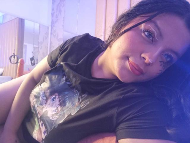 KonyBella, lying down, black t-shirt with cat graphic, blue eyes, short dark hair, slight smile, eyeliner, nose ring, soft purple and blue lighting, relaxed, slightly chubby.
