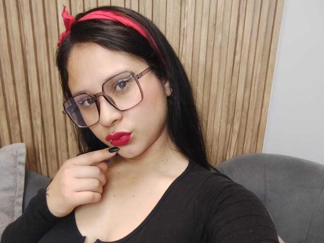 MaraVixen with glasses, red lipstick, black nails, black top, red headband, black hair, touching her lip, wood background, grey couch, medium tits, pale skin, blue eyes.