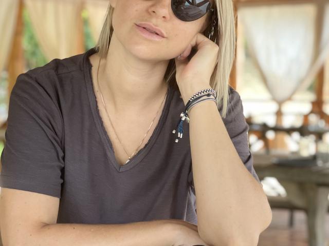 BelleLisaG with blonde hair, wearing dark sunglasses, a gray V-neck shirt, and a silver bracelet, resting her chin on her hand. She has a subtle, relaxed expression.