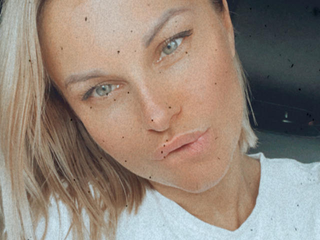 BelleLisaG has blonde hair, green eyes, and freckles, wearing a white shirt, with a pouty expression, looking slightly to the left in this selfie.