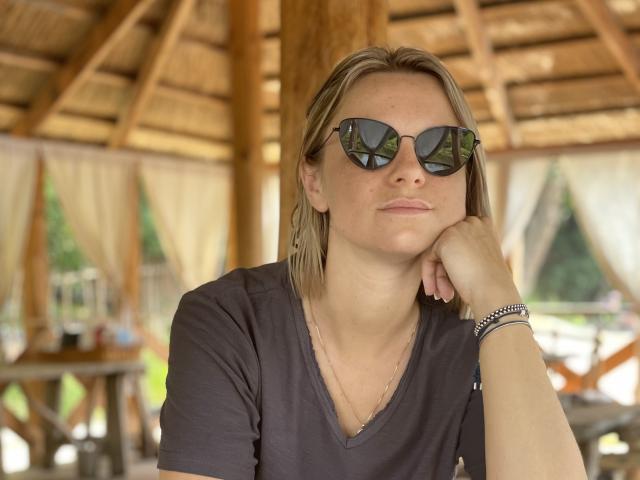 BelleLisaG with blonde hair, wearing reflective sunglasses, black v-neck shirt, and bracelets, resting her chin on her hand, looking relaxed.