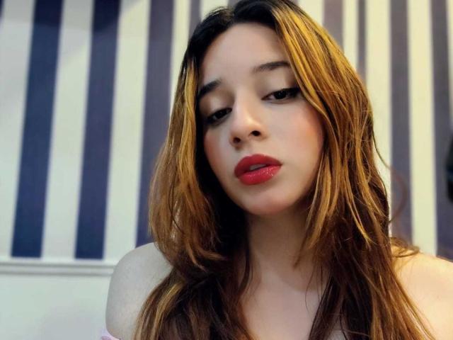 DaniVelasquez with long brown hair, red lipstick, and a slightly bored expression, showing off her bare shoulders. Her tits are partially visible. She's looking down at the camera.
