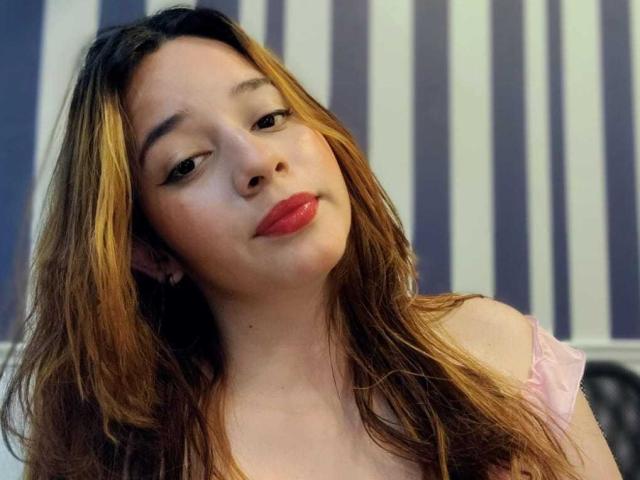 DaniVelasquez has light brown, wavy hair with blonde highlights, red lipstick, and a slightly tilted head, wearing a pink off-shoulder top, looking at the camera with a relaxed expression.