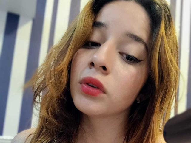 DaniVelasquez's face, with half-closed eyes, pink lips, and brown wavy hair, looking slightly bored or tired, in a selfie. Her tits aren't visible, just her face.