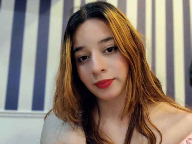 DaniVelasquez with long brown and blonde hair, red lipstick, and subtle makeup, looking at the camera with a slight smile. She has small tits and a cute face.