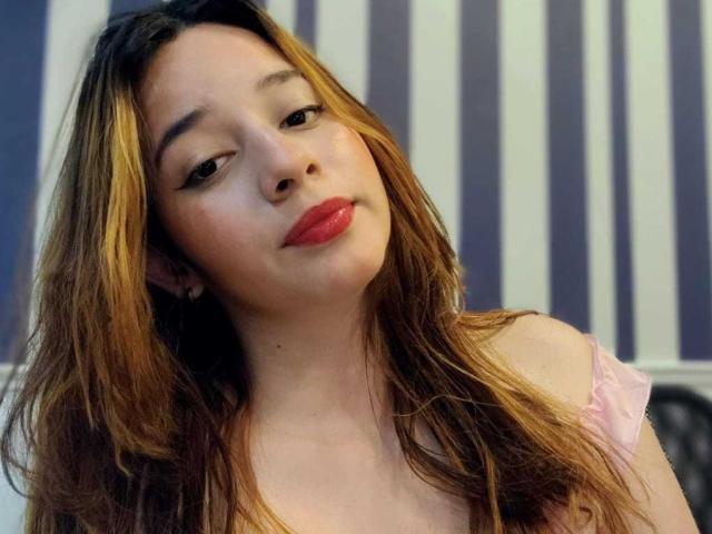 DaniVelasquez with light brown, wavy hair, red lipstick, and a slight smile, wearing a pink, off-shoulder top, looking to the side.