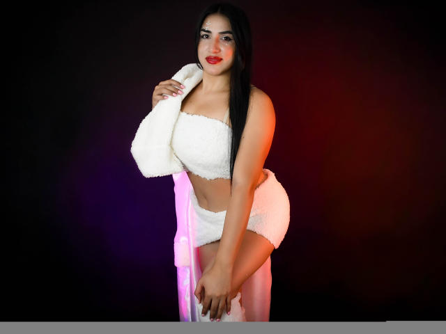 Alana Harris, avec ses longs cheveux noirs, portant un haut blanc duveteux sans bretelles et une jupe assortie, tenant une serviette blanche sur son épaule, avec du rouge à lèvres et un joli sourire, debout..