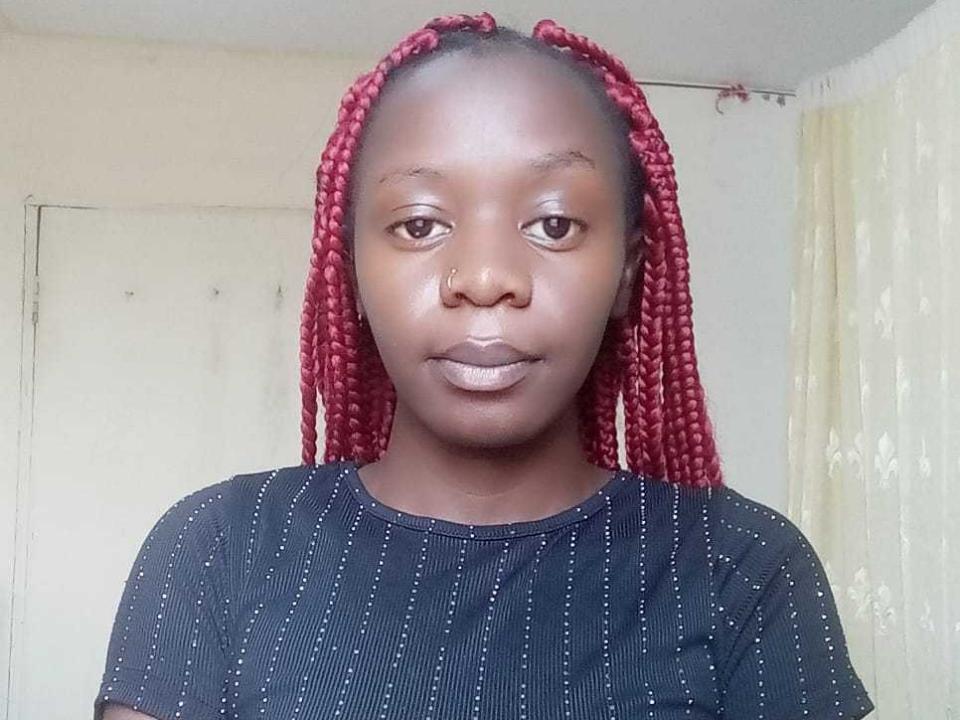 Slimthck with dark brown skin and red braids, wearing a black shirt with white lines, has a neutral expression and small nose, showing subtle makeup on her lips.