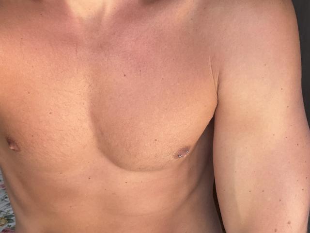 Joel69x's chest with a small nipple piercing on the right side, showing a tan line and slight chest hair, in a photograph.
