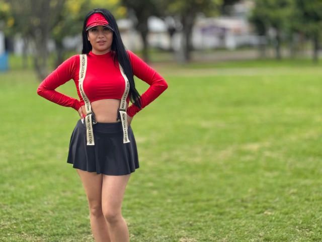 MiaHotmez with long black hair, red crop top, black skirt, suspenders, red visor, hands on hips, standing on green grass, showing her small tits and slim ass.