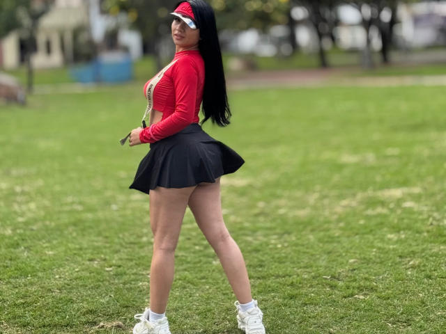 MiaHotmez with long black hair, red long-sleeve top, black pleated skirt, white sneakers, and a visor, showing her ass and small white waistband, standing on green grass.