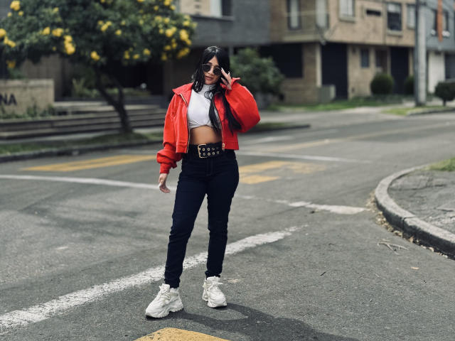 MiaHotmez in a red jacket, white crop top, black jeans, white sneakers, standing on a street, talking on her phone, with yellow flowers in the background.