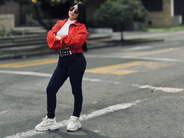 MiaHotmez with crossed arms, red jacket, white tee, black jeans, white sneakers, and a black belt, standing on an urban street, wearing dark sunglasses.