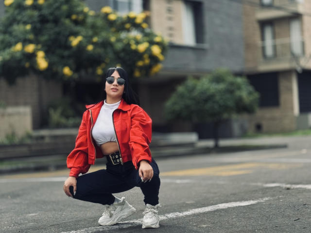 MiaHotmez is squatting, wearing a red jacket, white crop top, black pants, belt, and white sneakers. She has black hair, sunglasses, and a small nose ring.
