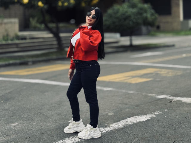 MiaHotmez with long black hair, red jacket, black pants, white sneakers, and sunglasses, standing in a street, smiling, hands on jacket, showing off her ass and tits.