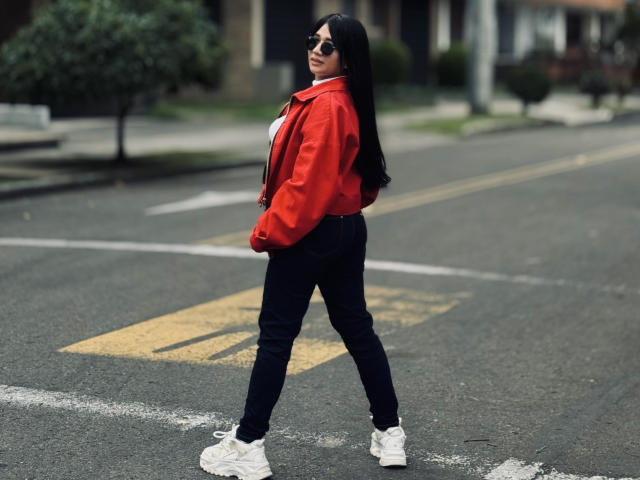 MiaHotmez with long black hair, red jacket, white shirt, black jeans, and white sneakers, standing on a street, looking over shoulder, wearing sunglasses.