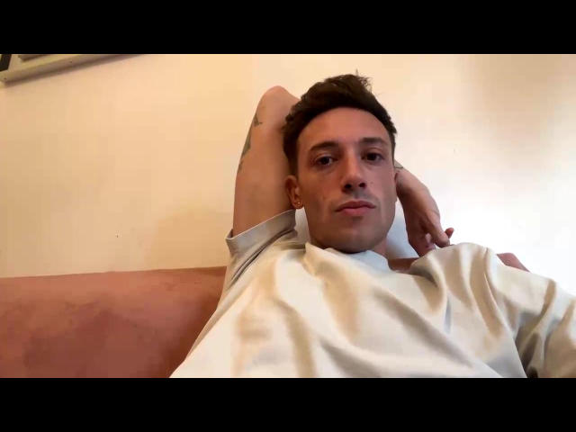 Joel69x, a dude with short brown hair, is lying on a pink couch, wearing a white tee, arms behind head, looking at the camera with a slight smile.