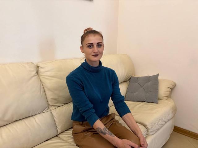 RileyWindow with red hair in a bun, wearing a teal turtleneck and brown pants, sitting on a cream leather couch, has dark lipstick and a tattoo on her right forearm.