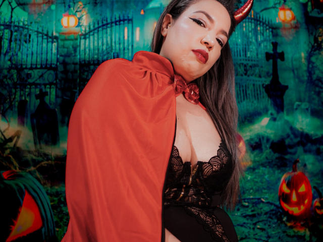 Mariehss with red devil horns, red cape, black lace lingerie, showing her big tits, long gray hair, and red lipstick, in a spooky Halloween-themed setting.