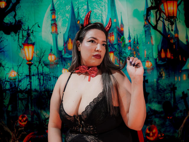 Mariehss with red devil horns, black lace dress, red ribbon choker, showing off her big tits, holding a finger to her mouth, dark lipstick, long brown hair.