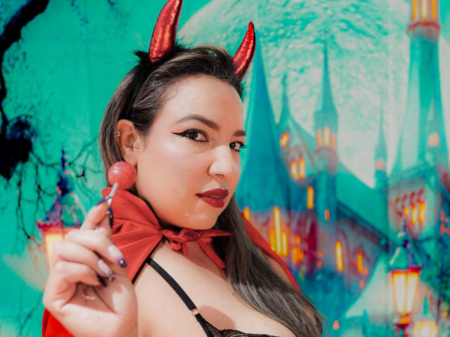 Mariehss with red devil horns, holding a lollipop, wearing a black bra, red bow, and headband, has dark makeup and glossy lips, showing off her tits.