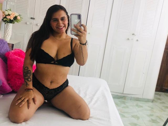 AmberDesire, kniend auf einem Bett, trägt einen schwarzen Spitzen-BH und Slips, zeigt ihre Brüste und Vulva, lächelt, hält ein Telefon, hat ein Tattoo am Arm, langes braunes Haar..