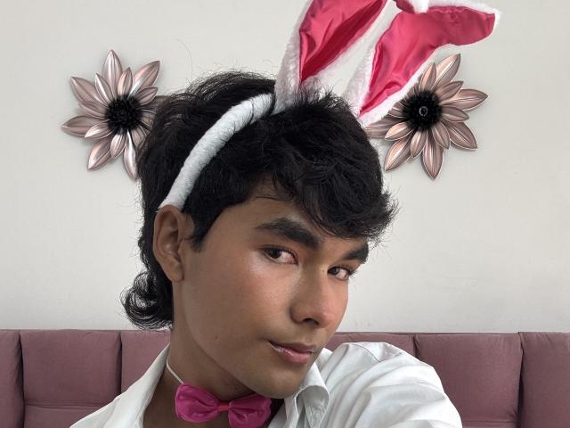 AngelDuzt has dark curly hair, brown skin, and wears pink bunny ears, a white headband, and a pink bowtie with a pink shirt, looking slightly to the side.