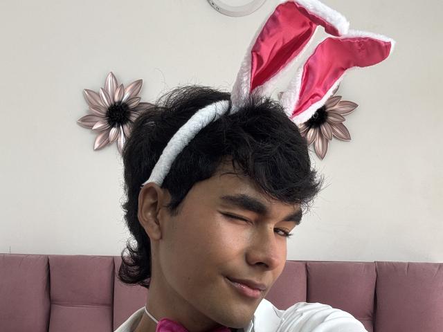 AngelDuzt has medium brown skin, black curly hair, and is wearing pink bunny ears with white trim, smiling with eyes closed, pink shirt, and pink upholstered background.