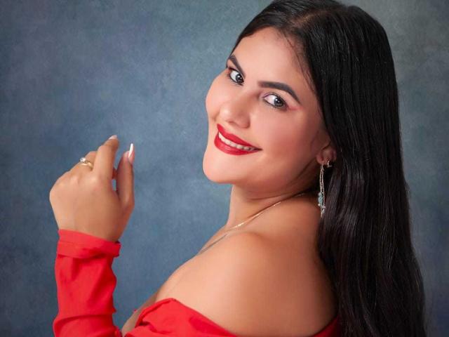 Sharlok with dark, straight hair, red lipstick, and red off-shoulder top, smiling at camera, holding hand up, showing ring, earrings, and necklace, with sexy, cute look.