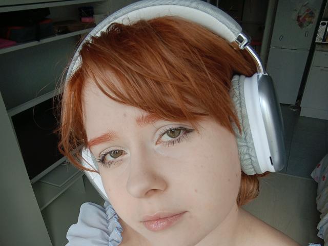 HoneyErica has red hair, light green eyes, and is wearing silver headphones with white cushions. Her skin is fair, and she's looking at the camera with a slight smile.